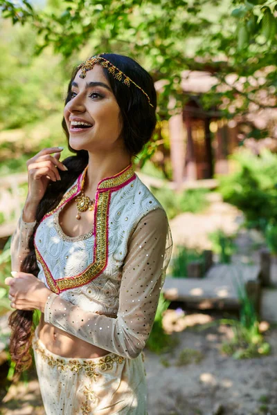 Элегантная и взволнованная индийская женщина в этнической одежде смотрит в сторону во время прогулки в парке — стоковое фото
