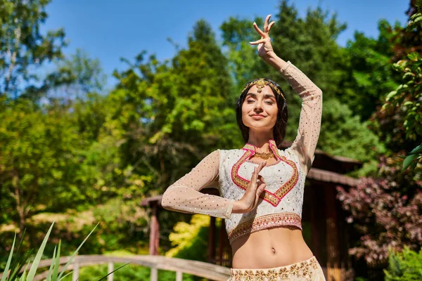 Летняя радость, молодая индианка в этнической одежде танцует с закрытыми глазами в солнечный день в парке — стоковое фото
