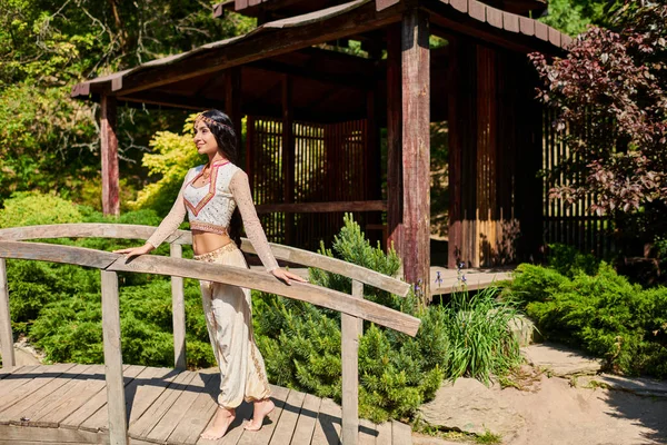 Heureuse femme indienne en vêtements authentiques debout sur le pont en bois près de l'alcôve dans le parc d'été — Photo de stock