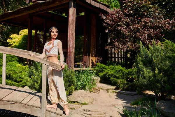 Mulher indiana morena em trajes autênticos vibrantes olhando para longe perto da ponte de madeira no parque de verão — Fotografia de Stock