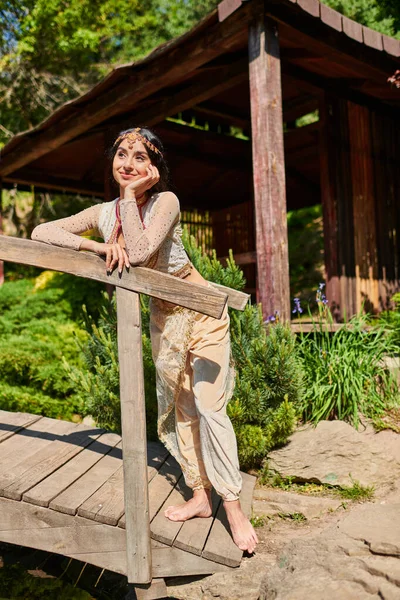 Mulher indiana feliz e sonhadora em traje tradicional olhando para longe na ponte de madeira no parque de verão — Fotografia de Stock