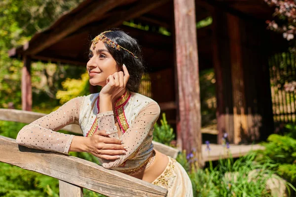 Parque de verano, soñadora mujer india de estilo auténtico sonriendo y mirando hacia otro lado en el puente de madera - foto de stock