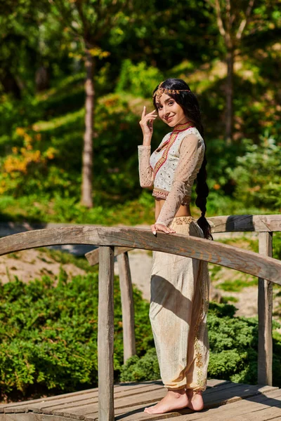 Donna indiana positiva e graziosa in abbigliamento etnico guardando la fotocamera sul ponte di legno nel parco soleggiato — Foto stock