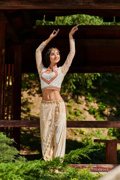Mujer india alegre y elegante en traje de estilo auténtico bailando y mirando hacia otro lado en el parque de verano - foto de stock