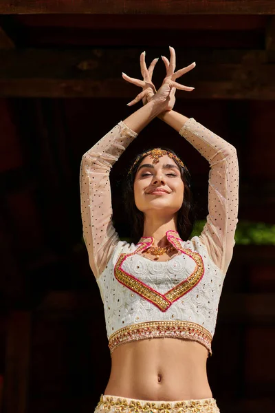Радісна молода індійська жінка в елегантному етнічному одязі танцює із закритими очима і піднімає руки на відкритому повітрі — стокове фото