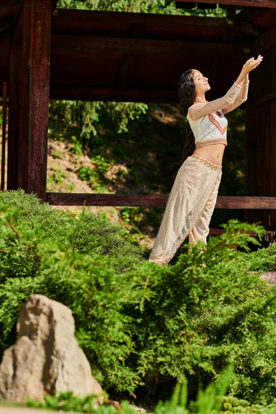 Traditional fashion, pleased indian woman in ethnic wear standing near green plants in summer park — Stock Photo
