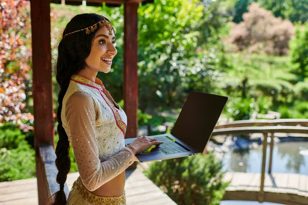 Летний парк, улыбающаяся индианка в стильной этнической одежде держит ноутбук с чистым экраном — стоковое фото
