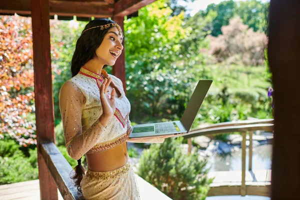 Joyful indian woman in traditional clothes waving hand during video call on laptop in summer park — Stock Photo