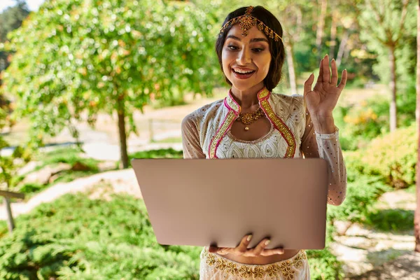 Joyful indian woman in traditional attire waving hand during video call on laptop in summer park — Stock Photo
