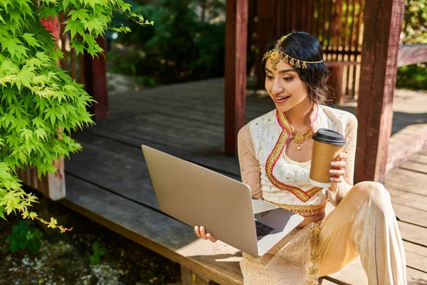 Femme indienne de style authentique avec tasse en papier et ordinateur portable assis dans l'alcôve dans le parc le jour de l'été — Photo de stock