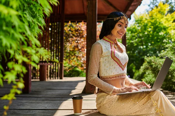 Sommerpark, glückliche Frau im ethnischen Stil tippt auf Laptop in der Nähe von Coffee to go in einer hölzernen Nische — Stockfoto