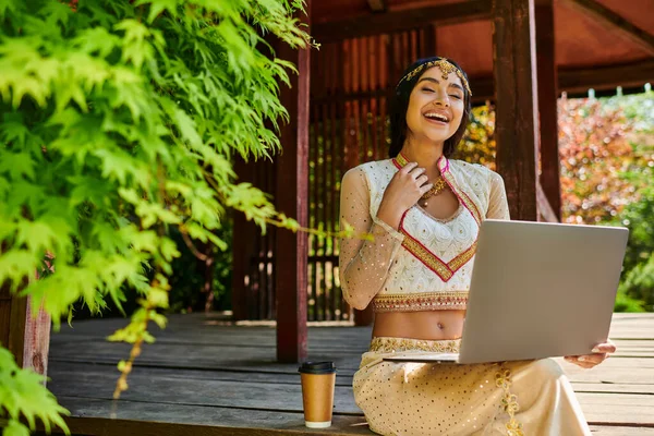 Mujer india riendo sentado con el ordenador portátil cerca del café para ir en la alcoba de madera en el día de verano - foto de stock