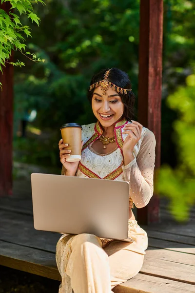 Mujer india alegre con café para ir mirando portátil en alcoba de madera en el parque en el día de verano - foto de stock