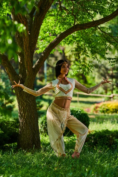 Elegante mujer india en vibrante traje étnico bailando bajo el árbol en el parque de verano, felicidad - foto de stock