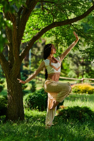 Молодая индианка в традиционной одежде танцует на зеленой лужайке под деревом в летнем парке — стоковое фото