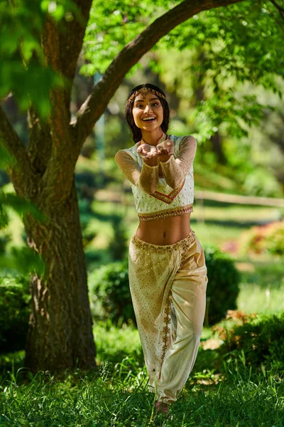 Réjouie, femme de style authentique regardant la caméra et pointant avec les mains sur la pelouse verte sous l'arbre — Photo de stock