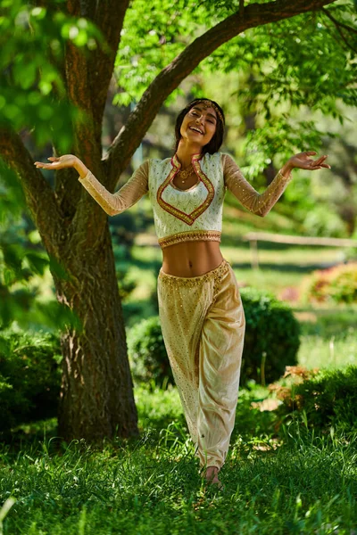 Carefree indian woman in vibrant traditional attire dancing in summer park on lawn under tree — Stock Photo