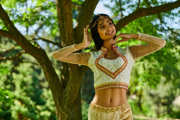 Mujer india elegante en ropa tradicional sonriendo y mirando hacia otro lado en el parque verde, ocio de verano - foto de stock