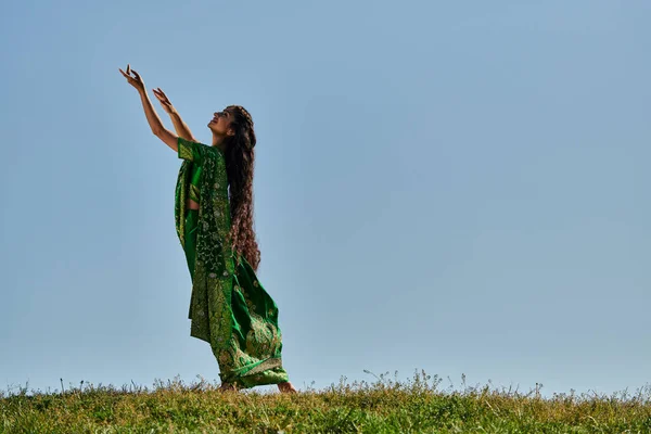 Día de verano, mujer india en ropa auténtica con las manos extendidas en el campo verde bajo el cielo azul - foto de stock
