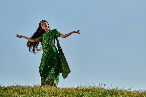 Femme indienne insouciante dans le sari traditionnel souriant à la caméra sur la pelouse verte sous le ciel bleu — Photo de stock