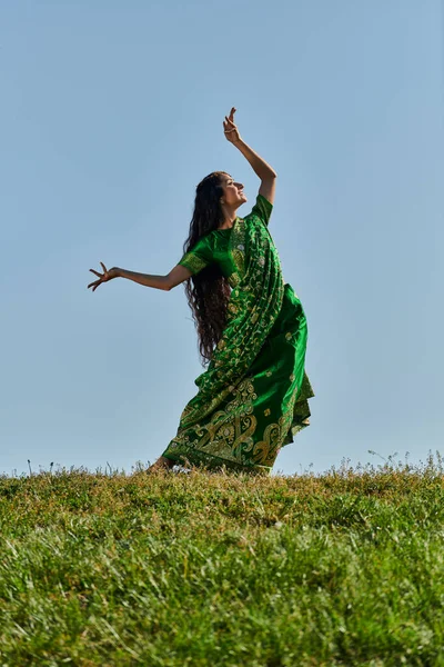 Young indian woman in sari dancing in green field under blue and clear sky, summer day — Stock Photo