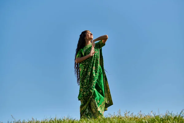 Disfrute del verano, campo verde, mujer india en ropa étnica sonriendo con los ojos cerrados bajo el cielo azul - foto de stock