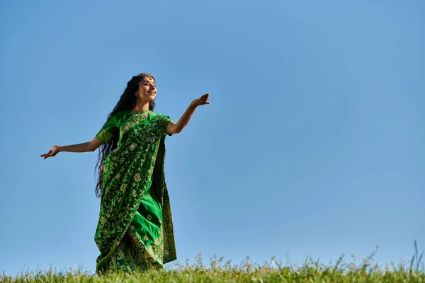 Элегантная индийская женщина в традиционном сари танцует на зеленом лугу под голубым летним небом — стоковое фото