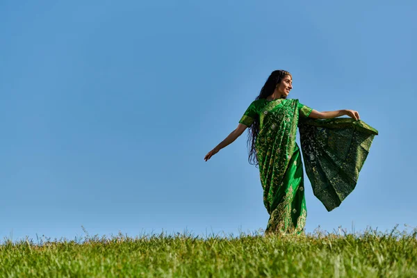 Día de verano, mujer india despreocupada en ropa auténtica caminando en el campo verde bajo el cielo azul - foto de stock