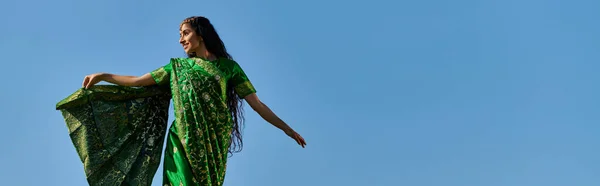 Ocio de verano, mujer india en sari sonriendo y mirando hacia otro lado bajo el cielo azul sin nubes, bandera - foto de stock