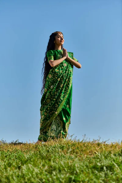 Mulher indiana feliz em sari com as mãos de oração e olhos fechados no gramado sob o céu azul, dia de verão — Fotografia de Stock