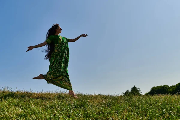 Вільна розумна індійська жінка в сарі працює на зеленому лузі під блакитним небом, щасливе літо — стокове фото