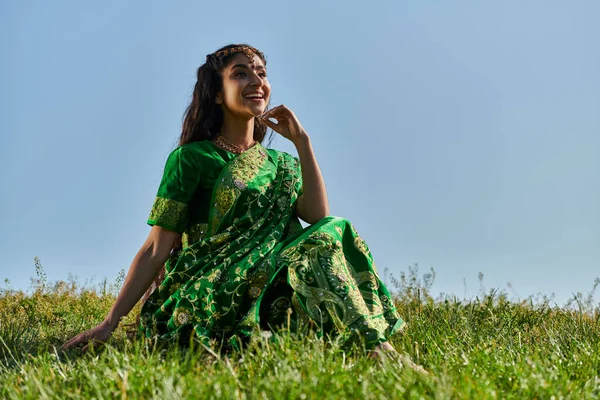 Позитивная молодая индийская женщина в зеленом сари сидит на травянистом холме с голубым небом на заднем плане — стоковое фото