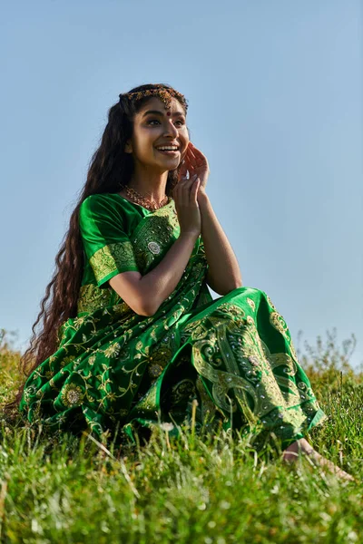 Fröhliche indische Frau in Sari und matha patti sitzt auf einem grasbewachsenen Hügel mit blauem Himmel auf dem Hintergrund — Stockfoto