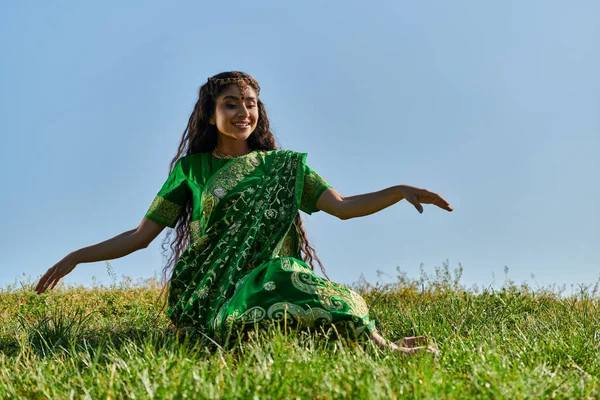 Sonriente joven india mujer en verde sari sentado en la colina cubierta de hierba con el cielo azul en el fondo - foto de stock