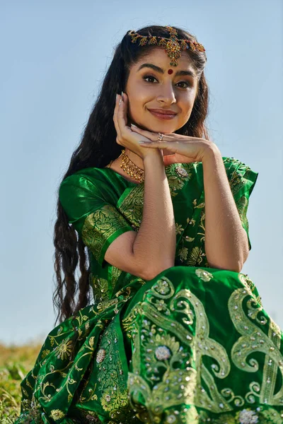 Sonriente mujer india en sari y matha patti mirando la cámara en la colina cubierta de hierba y el cielo en el fondo - foto de stock
