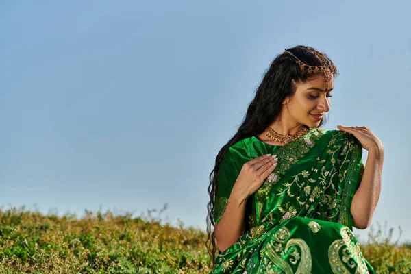 Mujer india joven con Matha Patti tocando sari verde en la colina con el cielo azul en el fondo - foto de stock