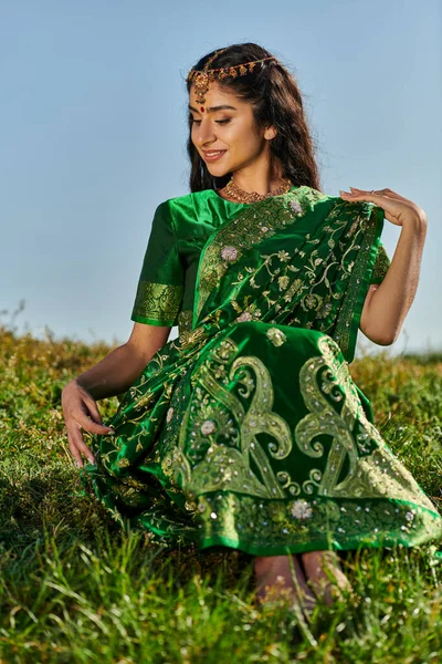 Sonriente joven india mujer tocando verde sari mientras está sentado en la colina cubierta de hierba con el cielo en el fondo - foto de stock