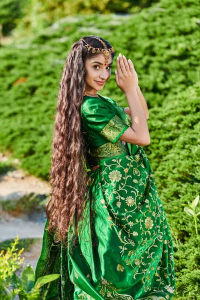 Lächelnde langhaarige Indianerin im grünen Sari posiert in der Nähe von Pflanzen im Park im Freien — Stockfoto