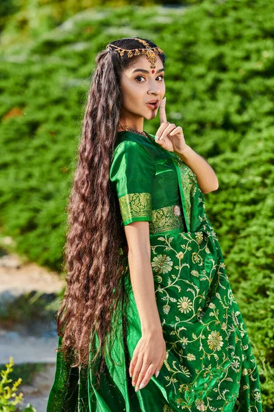 Jovem indiana mulher no sari mostrando gesto secreto e olhando para a câmera perto de plantas no parque — Fotografia de Stock