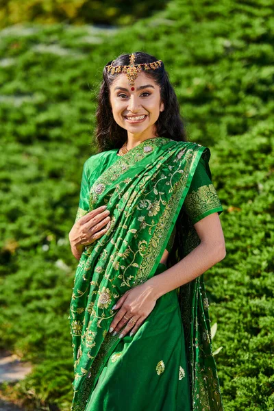 Retrato de jovem mulher indiana alegre em sari tradicional e matha patti olhando para câmera no parque — Fotografia de Stock