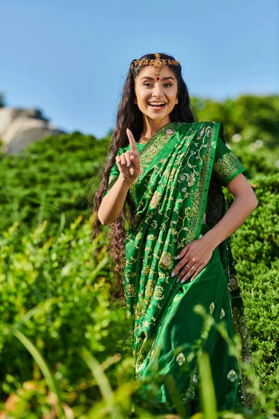 Fröhliche junge Indianerin im Sari zeigt mit dem Finger, während sie in der Nähe von Pflanzen im Park steht — Stockfoto
