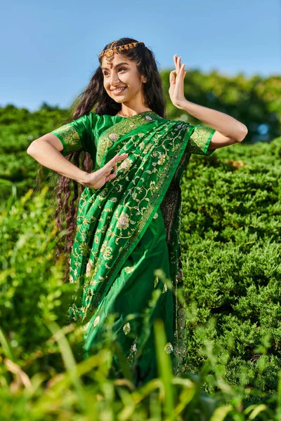 Jovem mulher indiana despreocupada em sari tradicional dançando enquanto está perto de plantas no parque — Fotografia de Stock