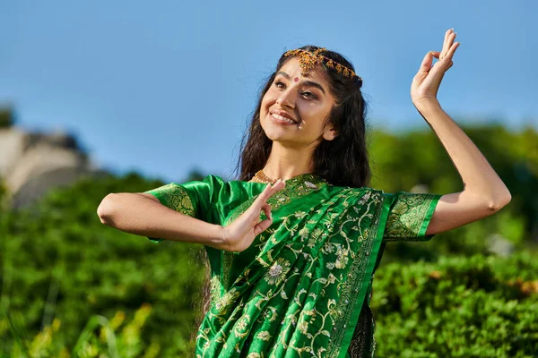Bonita jovem mulher indiana em sari tradicional dançando no parque borrado com céu azul no fundo — Fotografia de Stock