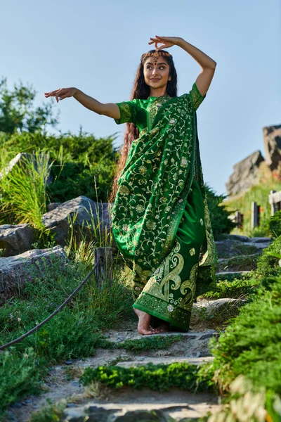 Mujer india joven con estilo en sari verde de pie en las escaleras de piedra en el parque de verano - foto de stock