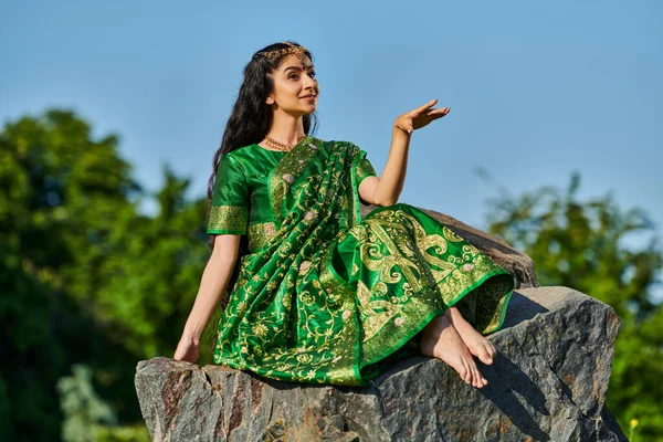 Sonriente y elegante mujer india descalza en sari sentado en piedra con el cielo azul en el fondo - foto de stock