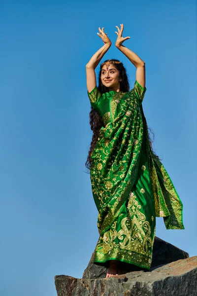 Alegre joven india mujer en verde sari posando sobre piedra con cielo azul sobre fondo - foto de stock