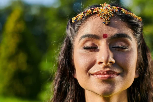 Retrato de mujer india sonriente con bindi y matha patti de pie con los ojos cerrados al aire libre - foto de stock