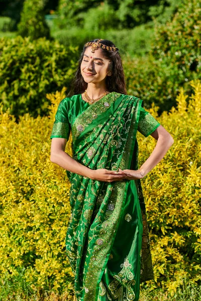 Elegante e sorridente jovem indiana mulher em sari posando em arbustos no parque ao ar livre — Fotografia de Stock