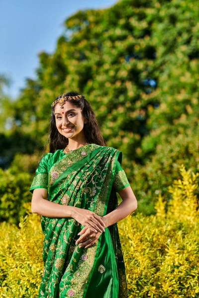 Mujer india bonita y sonriente en sari mirando a la cámara mientras posa cerca de las plantas en el parque - foto de stock