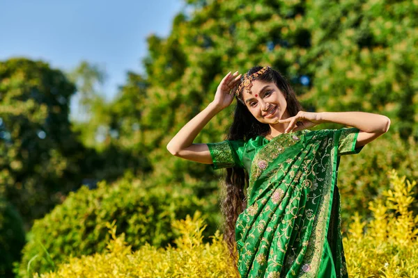 Позитивная молодая индийская женщина в сари и бинди смотрит в камеру рядом с растениями в парке — стоковое фото